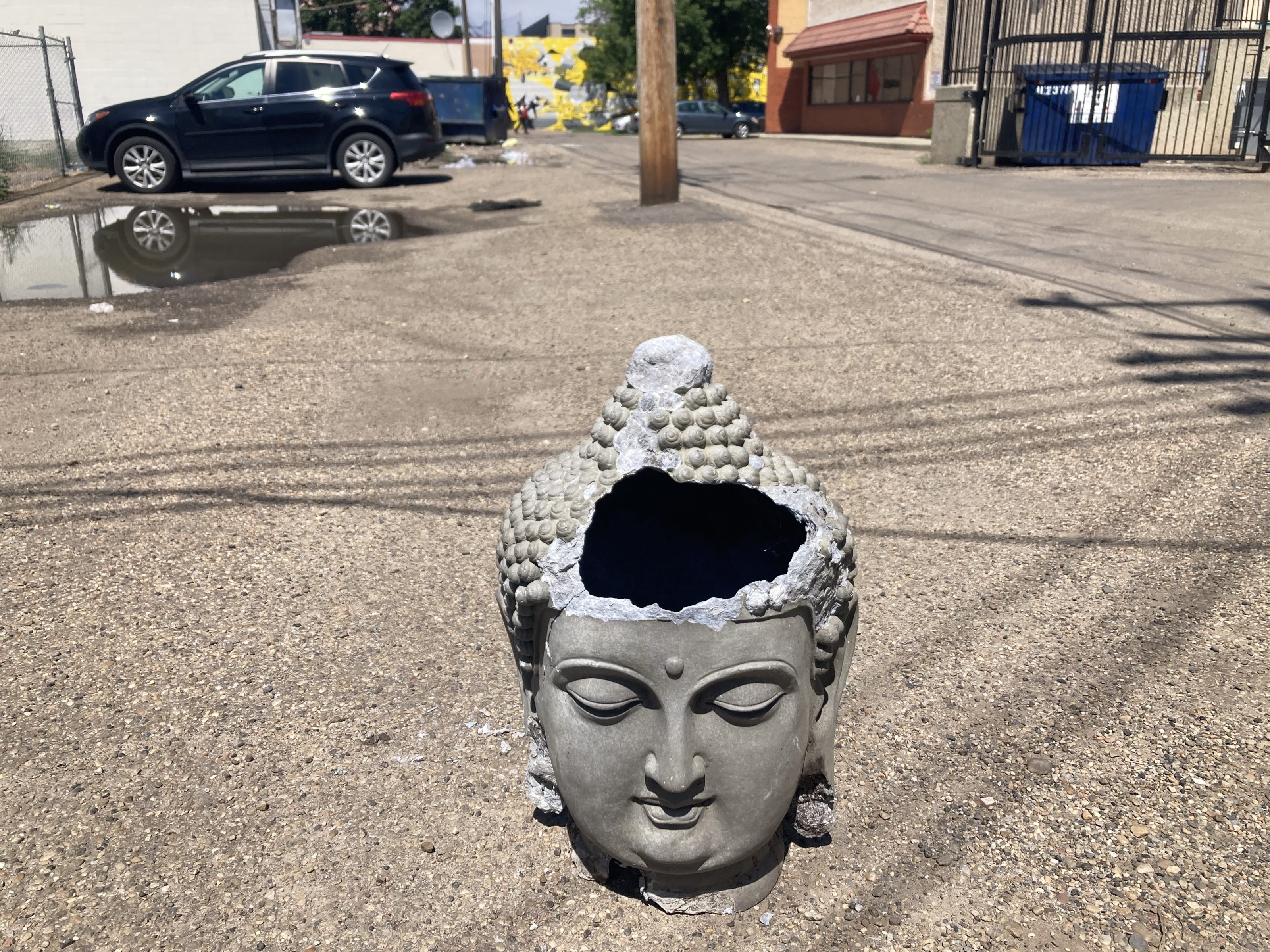 A broken statue of the Buddha's head exposing the darkness within it sits in the middle of a sunlit alley amid the shadows of wires, a dumpster, and a large puddle.  