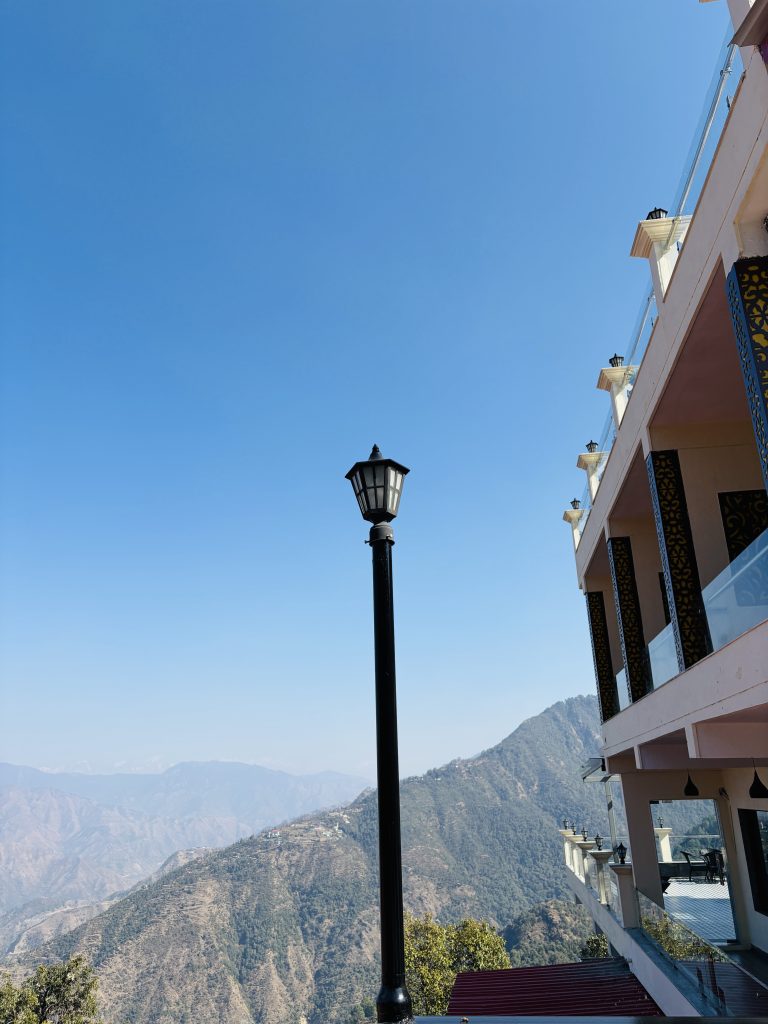 A scenic view featuring a clear blue sky, a lamp in the foreground, and a mountain landscape in the background. On the right, a building with multiple balconies and decorative elements is visible.