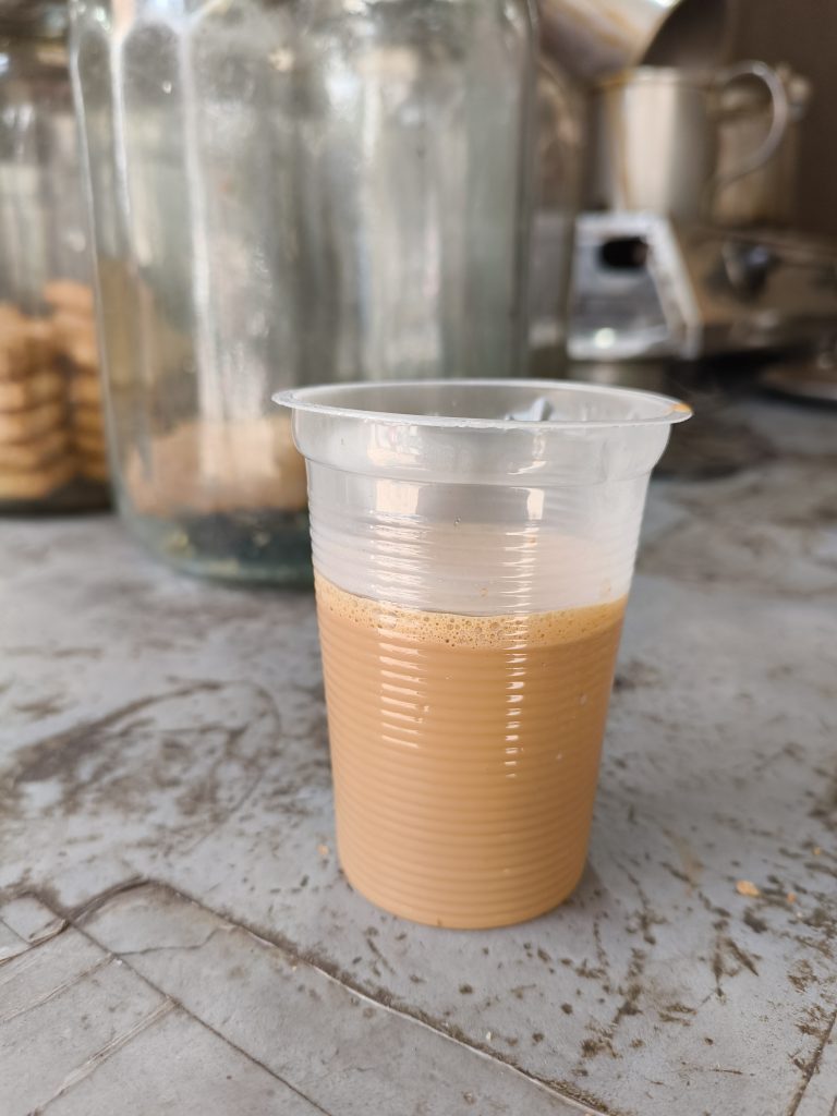 A plastic cup filled with creamy-looking chai placed on a textured surface, with blurred glass containers and metal kitchenware in the background.