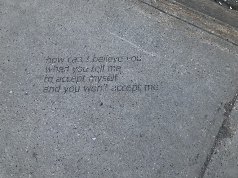 A segment of a sidewalk with cigarette butts and the engraved words of a homeless person: how can I believe you when you tell me to accept myself and you won’t accept me.