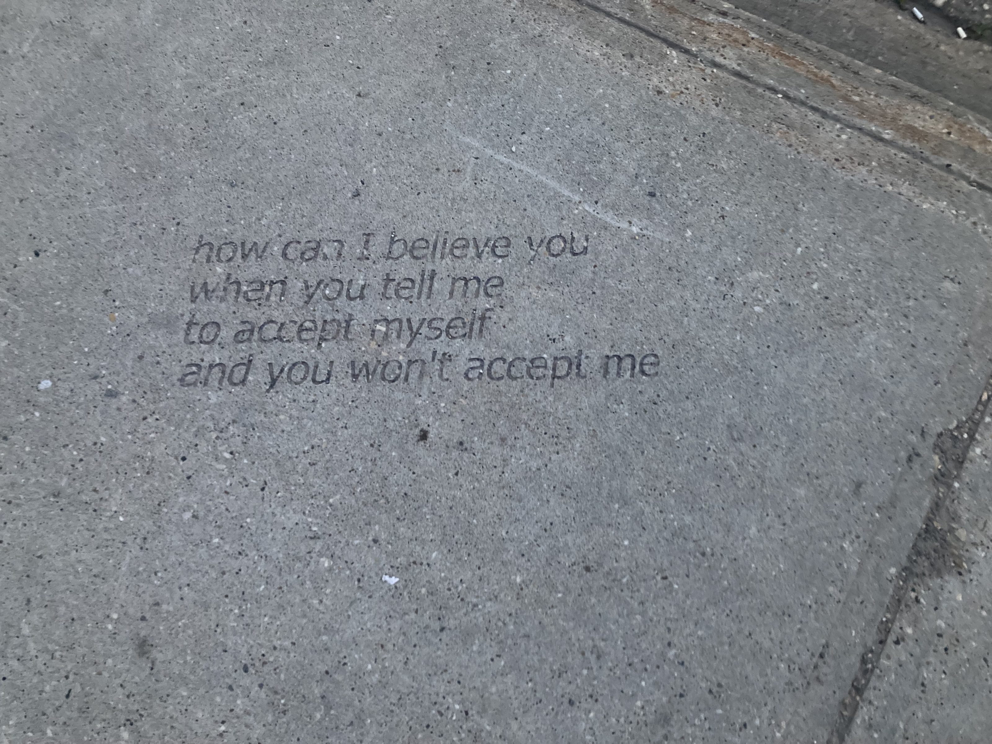 A segment of a sidewalk with cigarette butts and the engraved words of a homeless person: how can I believe you when you tell me to accept myself and you won't accept me.