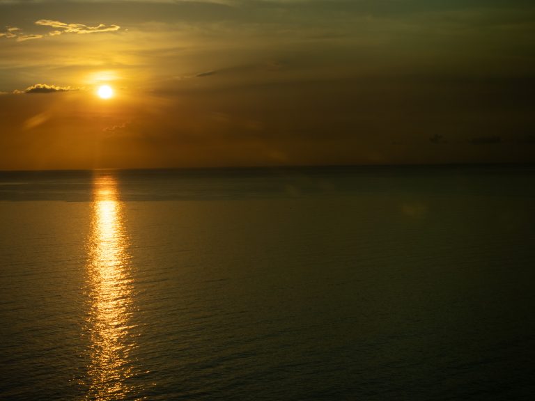 A serene sunset over the ocean with the sun low in the sky, casting a golden reflection on the calm water and clouds scattered across the horizon.