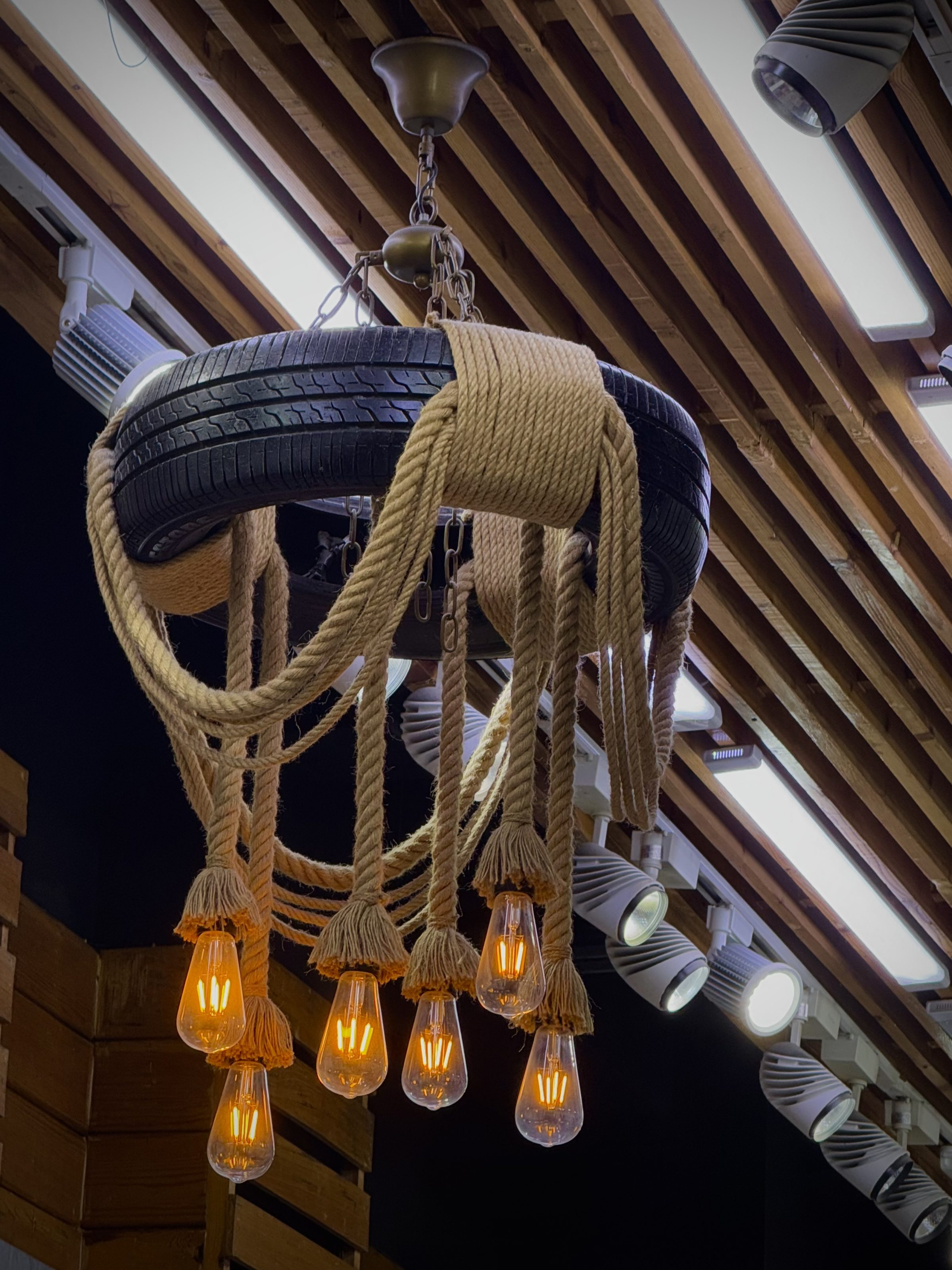 A chandelier made from an upcycled tire hangs from the ceiling, wrapped with thick rope. Several glowing Edison-style bulbs hang from the rope, creating a warm, rustic ambiance. The ceiling features wooden slats and modern lighting fixtures in the background.