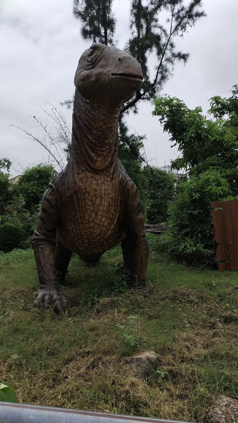 Life-sized dinosaur model in a lush green park setting, surrounded by trees and grass, under a cloudy sky.