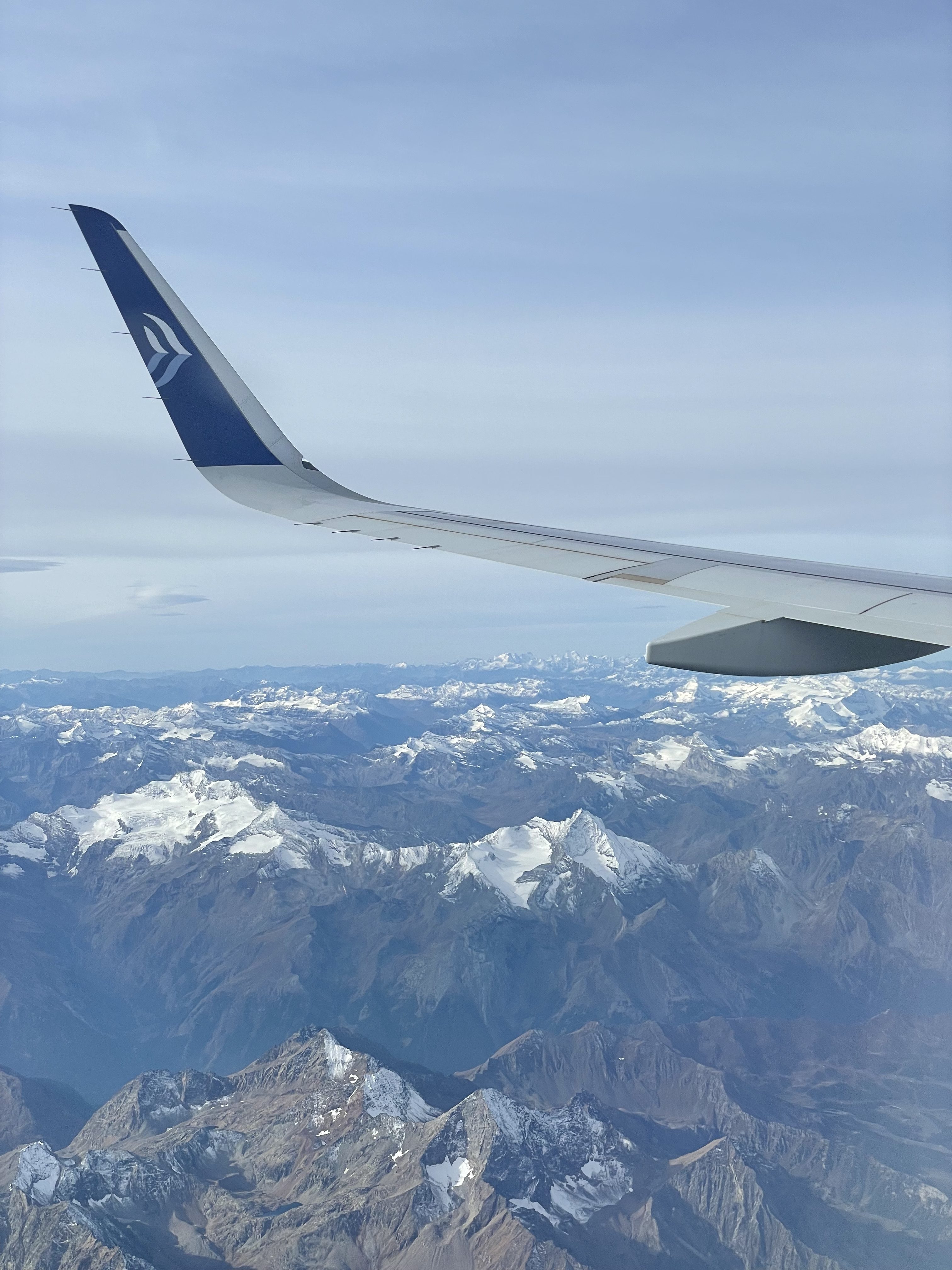 View from an airplane window showing a wing and the mountainous terrain below, covered with snow-capped peaks under a clear sky.