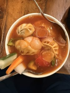 A bowl of soup with dumplings, sliced onions, and bell peppers in a tomato-based broth, garnished with raw cucumber, carrot, and radish sticks, with a spoon placed on the edge.