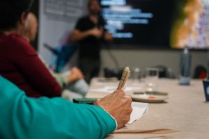 A person holding a pen is taking notes at a table during a presentation. Other people are seated around the table, and a blurred presenter is visible in front of a screen with text.