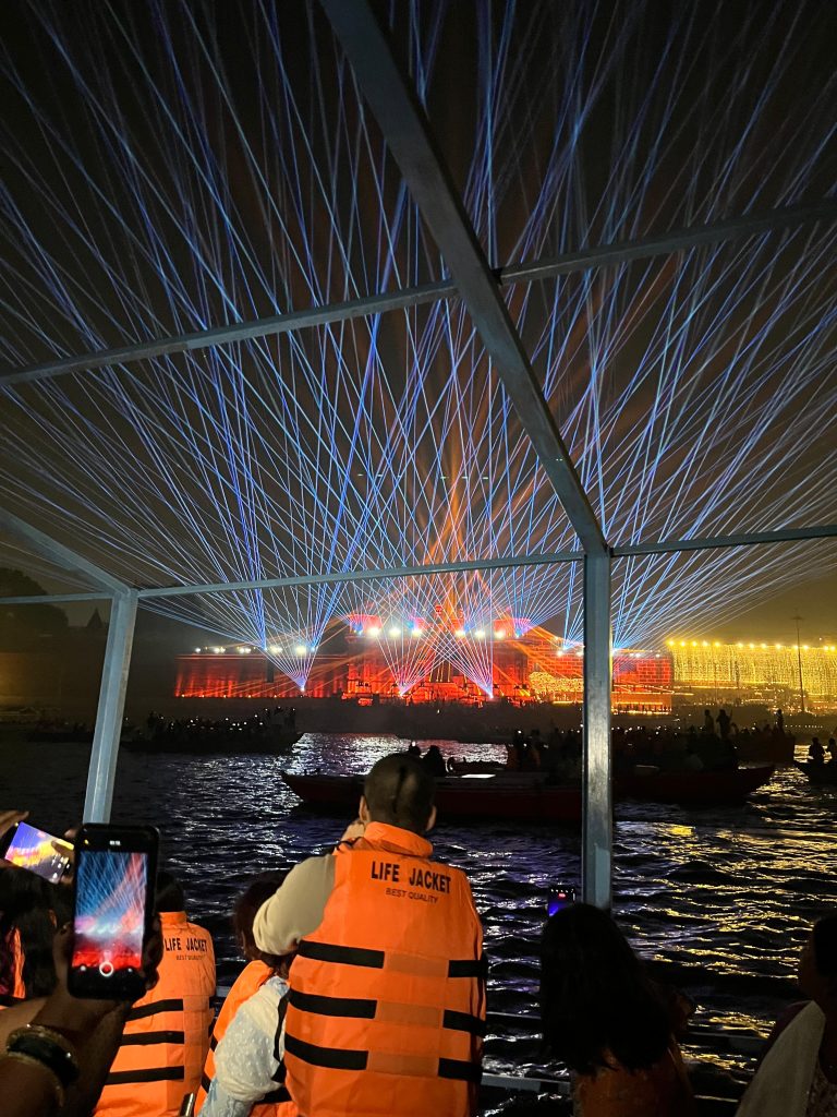 People wearing orange life jackets are on a boat watching a colorful laser light show projected on a building across the water.