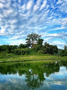 A tranquil landscape featuring a lush green hill topped with trees, reflected in the calm waters of a lake. The blue sky is filled with scattered white clouds.