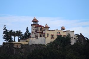 A historic building with domed towers and arches, set atop a rocky hill with surrounding greenery and a clear blue sky background.