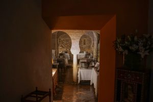 A dimly lit, cozy restaurant interior featuring arched brick walls and columns. The room is decorated with neatly arranged tables covered with white tablecloths and set with glassware