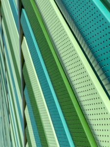 The image showcases a close-up of a modern architectural facade featuring diagonal, perforated metal panels in shades of green. The panels create a dynamic, textured pattern with alternating colors and small circular perforations.