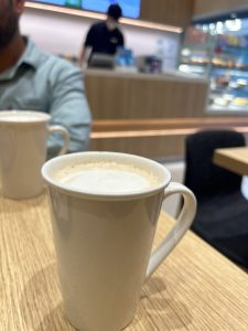 A close-up of a white coffee mug on a wooden table in a cafe, with a blurred person in the background at the counter and a display case to the side.