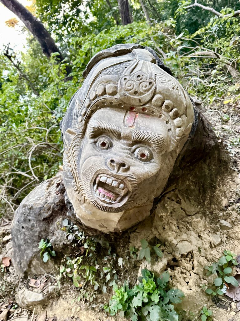 A stone sculpture of a fierce, snarling face with intricate carvings and decorations, partially embedded in the ground, surrounded by greenery and forest vegetation.