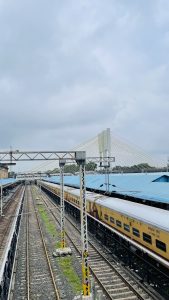 A railway station with a parked train on the right side and empty tracks on the left. The station features blue-roofed platforms and overhead electric wires