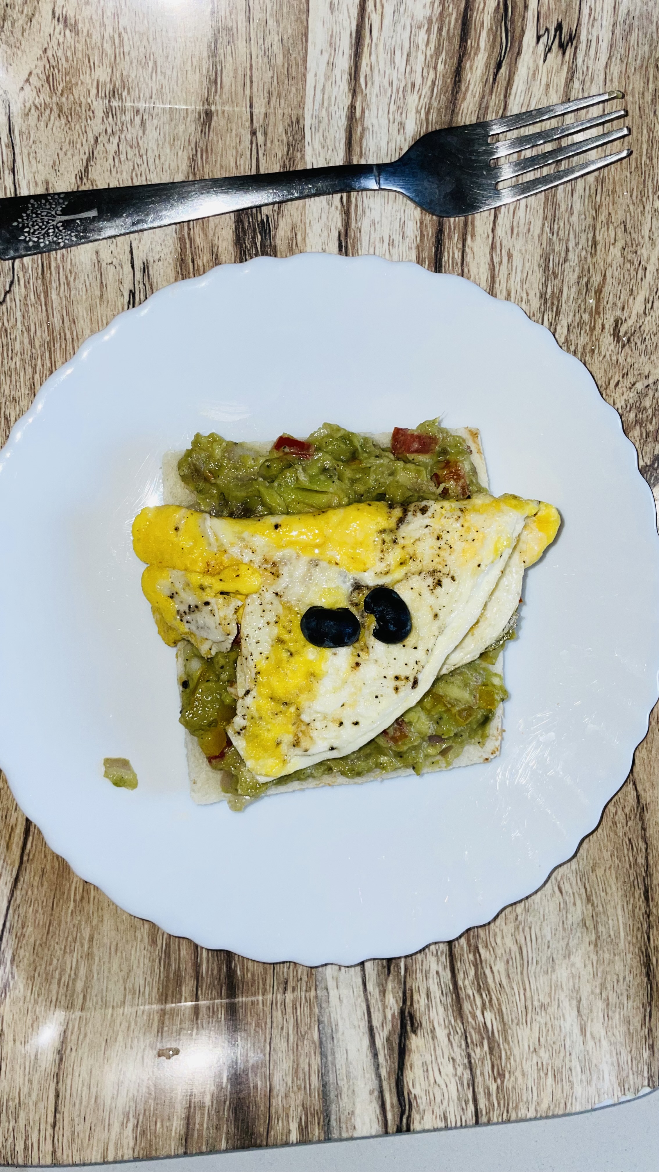 A white plate with a slice of toasted bread topped with smashed avocado, a folded omelet, and black olive slices, placed on a wooden table. A fork is positioned above the plate.