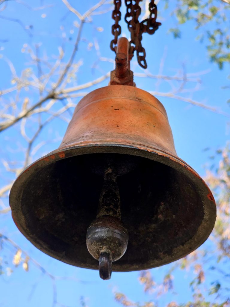 A rusty, metal bell hanging from a chain with a clear blue sky and tree branches in the background.