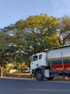 A white tanker truck is parked on the side of a road, with large green trees in the background under a clear blue sky.