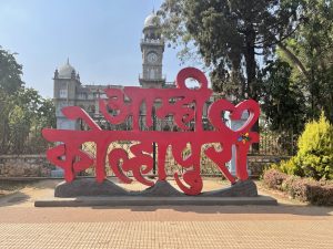 A large red sculpture with the text in a decorative script is in the foreground. Behind it, there is a historical building with domes and a clock tower, surrounded by trees and greenery. The sky is clear and blue.