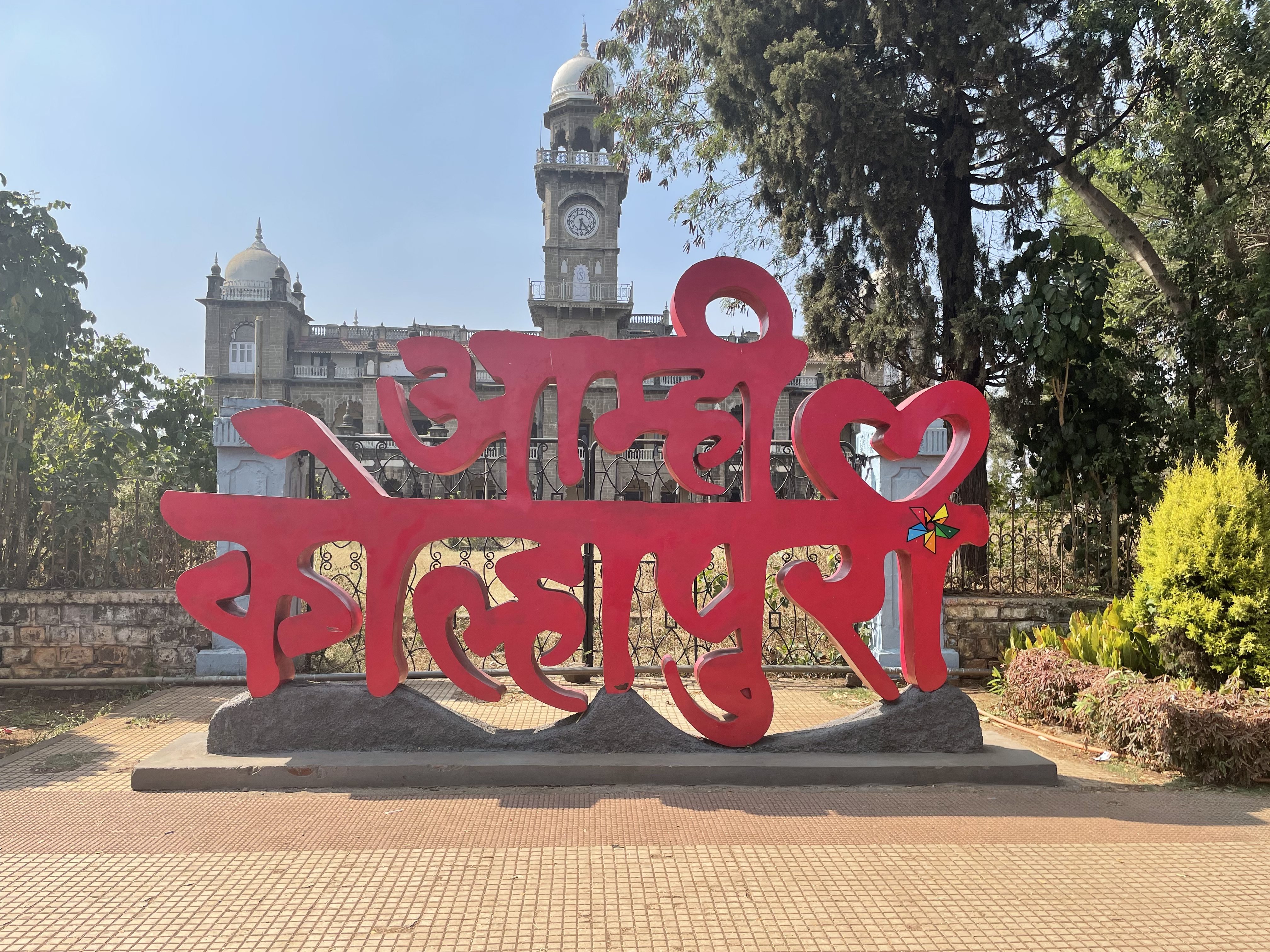 A large red sculpture with the text in a decorative script is in the foreground. Behind it, there is a historical building with domes and a clock tower, surrounded by trees and greenery. The sky is clear and blue.