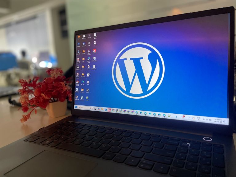 A laptop on a desk with a large WordPress logo displayed on the screen. To the left of the laptop is a small potted plant with red and pink flowers. In the background, there’s a blurred office environment.