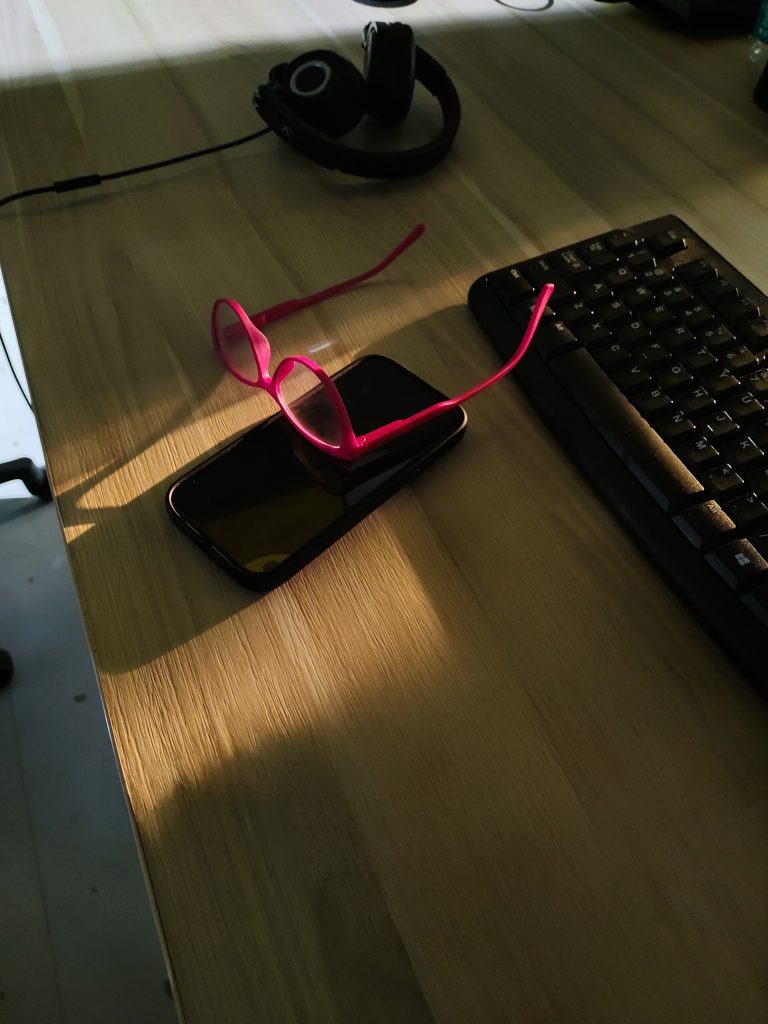 A wooden desk with a black keyboard, a pair of pink eyeglasses resting on a smartphone, and a set of black headphones nearby.