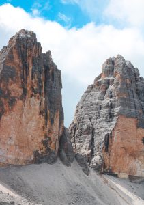 Two towering rock formations stand against a backdrop of a blue sky with scattered clouds. The rugged cliffs have a reddish and grayish hue, with visibly stratified layers. Italian Dolomites 