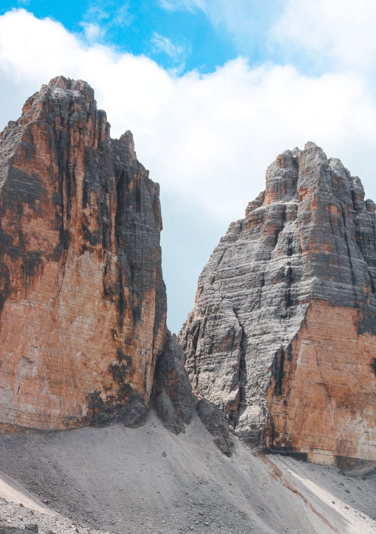 Two towering rock formations stand against a backdrop of a blue sky with scattered clouds. The rugged cliffs have a reddish and grayish hue, with visibly stratified layers. Italian Dolomites