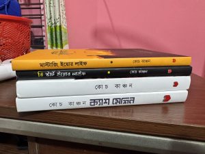 A stack of four Bengali books on a wooden desk, with yellow, black and white covers. A red basket and floral curtain are visible in the background against a pink wall.