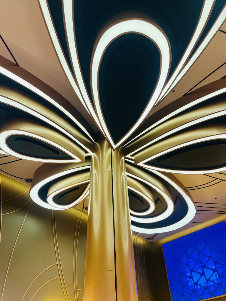 Abstract architectural design featuring a large, central gold pillar extending to a ceiling composed of curved, illuminated shapes. The wall is adorned with geometric patterns and a section in blue.