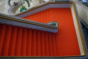 Aerial view of a red-carpeted staircase with gray concrete edges, featuring a unique, angular design. In the background, a small kid is using the polished handrail as a slide.