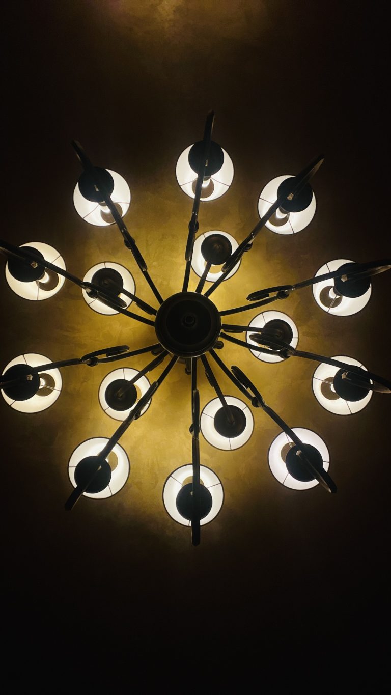 A close-up view of a circular chandelier with multiple arms and lit round bulbs, casting a warm glow against a dark ceiling.