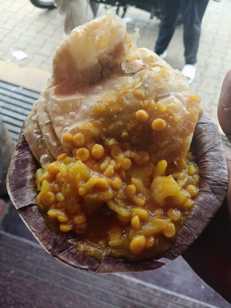 A close-up of a traditional Indian street food dish served on a leaf plate, featuring a flaky, layered puri accompanied by a yellow lentil curry.