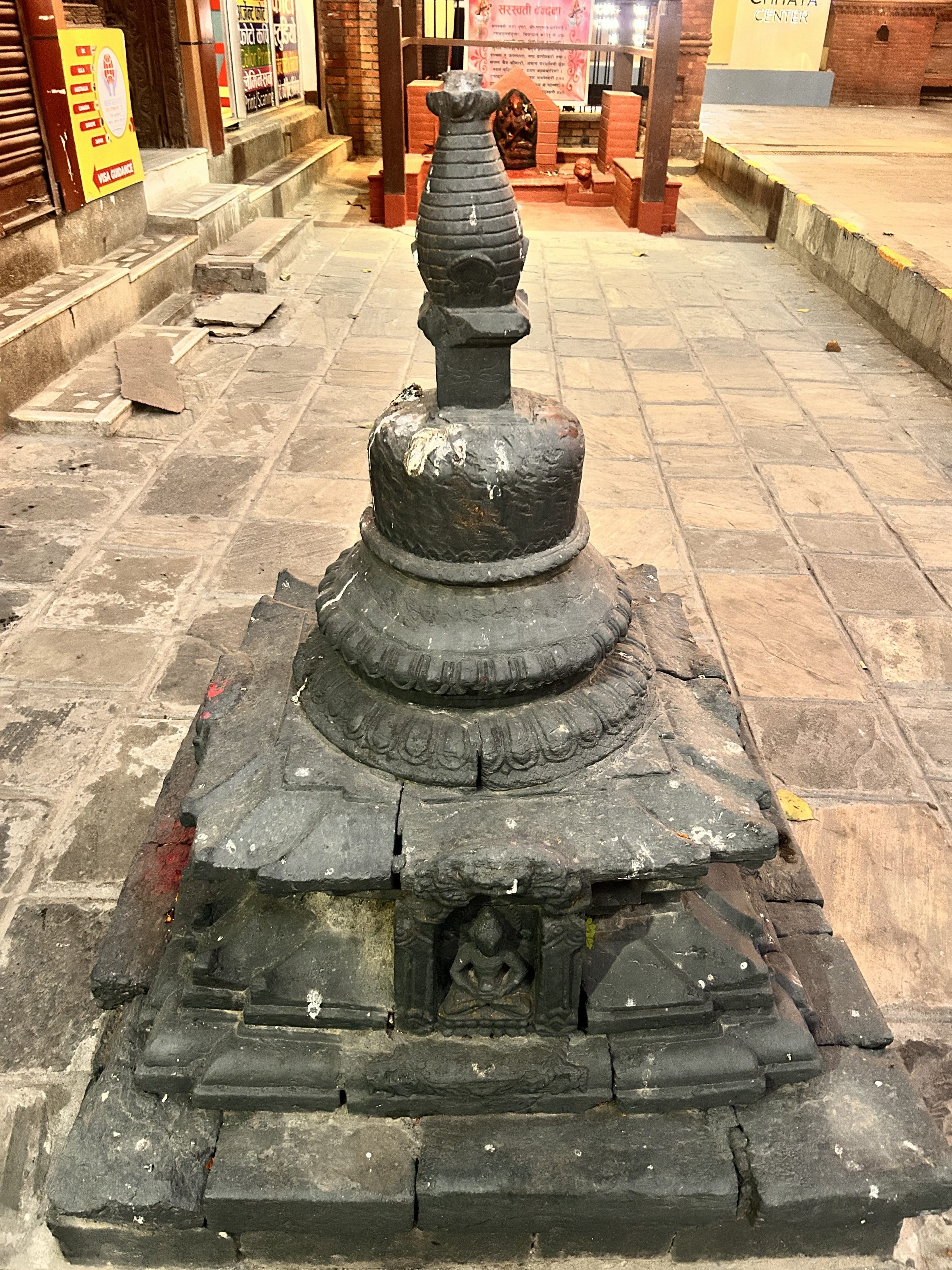 A small stone stupa with intricate carvings on its base, located on a paved stone path. The background features brick structures and some signage.