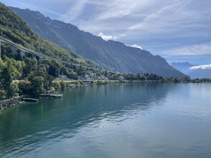 A serene lake with clear, calm water reflecting the sky, surrounded by lush green hills and mountains under a partly cloudy blue sky