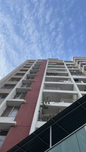 Upward view of a tall residential building with white and red sections, balconies with railings, and a blue sky with scattered clouds in the background.
