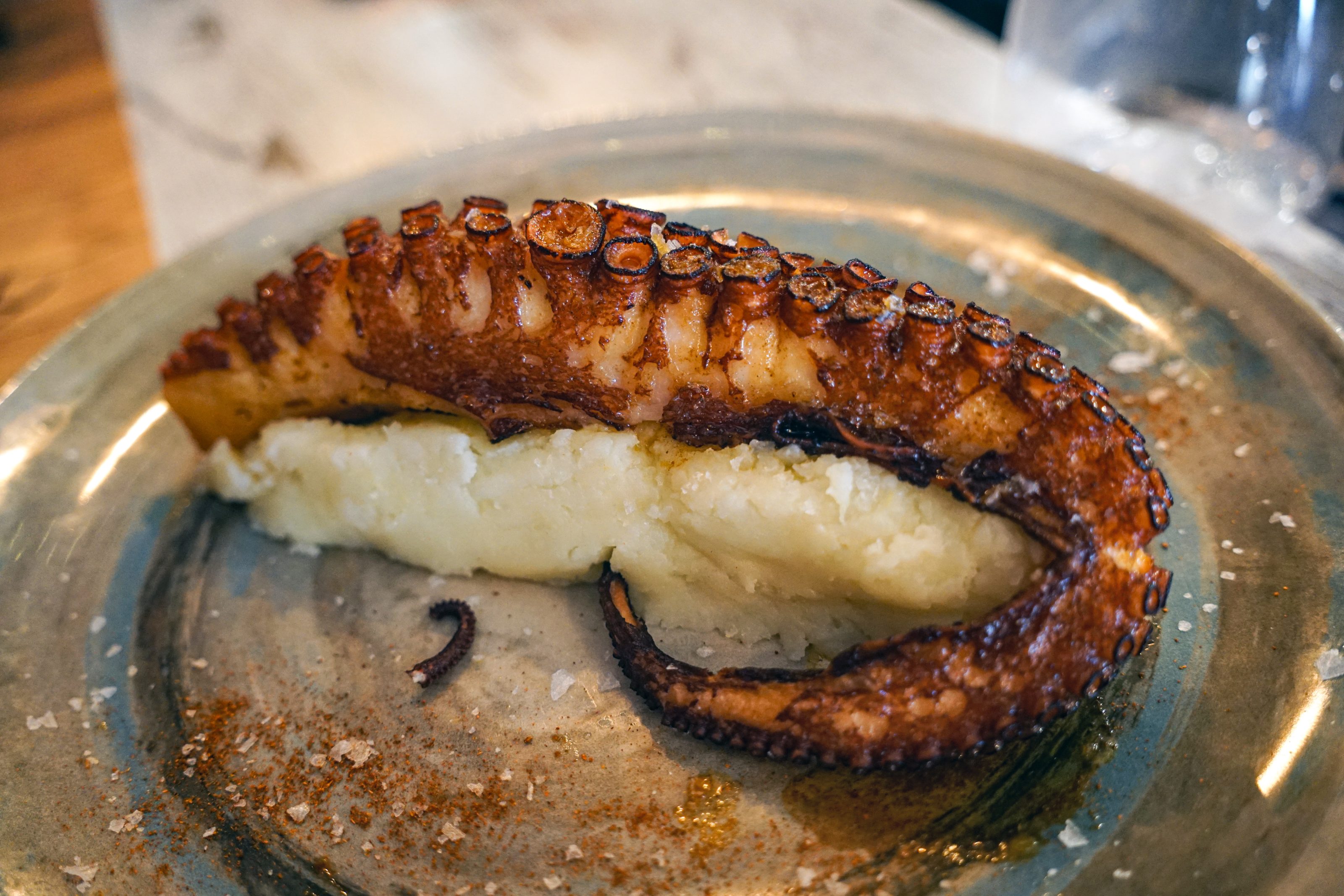 A grilled octopus tentacle served on a bed of mashed potatoes, garnished with flakes of sea salt and a sprinkle of spices on a ceramic plate.