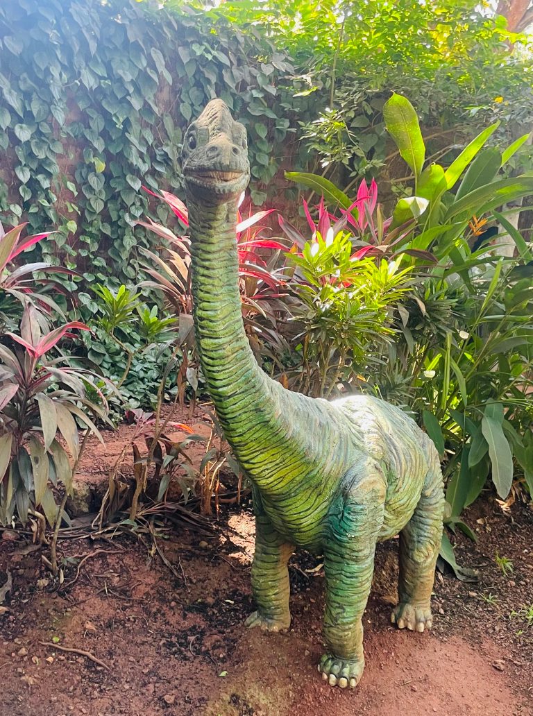 A sculpture of a green dinosaur with a long neck and a smiling face, surrounded by lush green foliage and colorful plants in a garden setting.