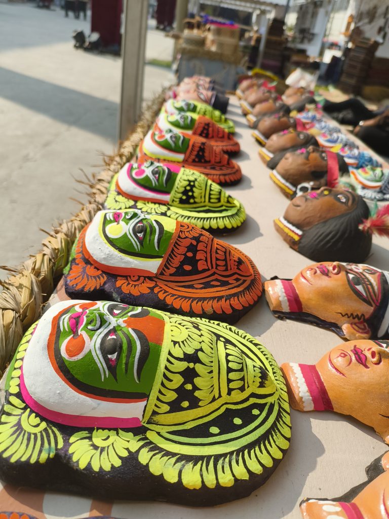 Colorful clay masks with intricate designs are displayed on a table at an outdoor market, showcasing vibrant greens, oranges, and other hues.