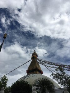 
A large Bouddha stupa with eyes painted on it under a cloudy sky, adorned with colorful prayer flags extending from the spire.