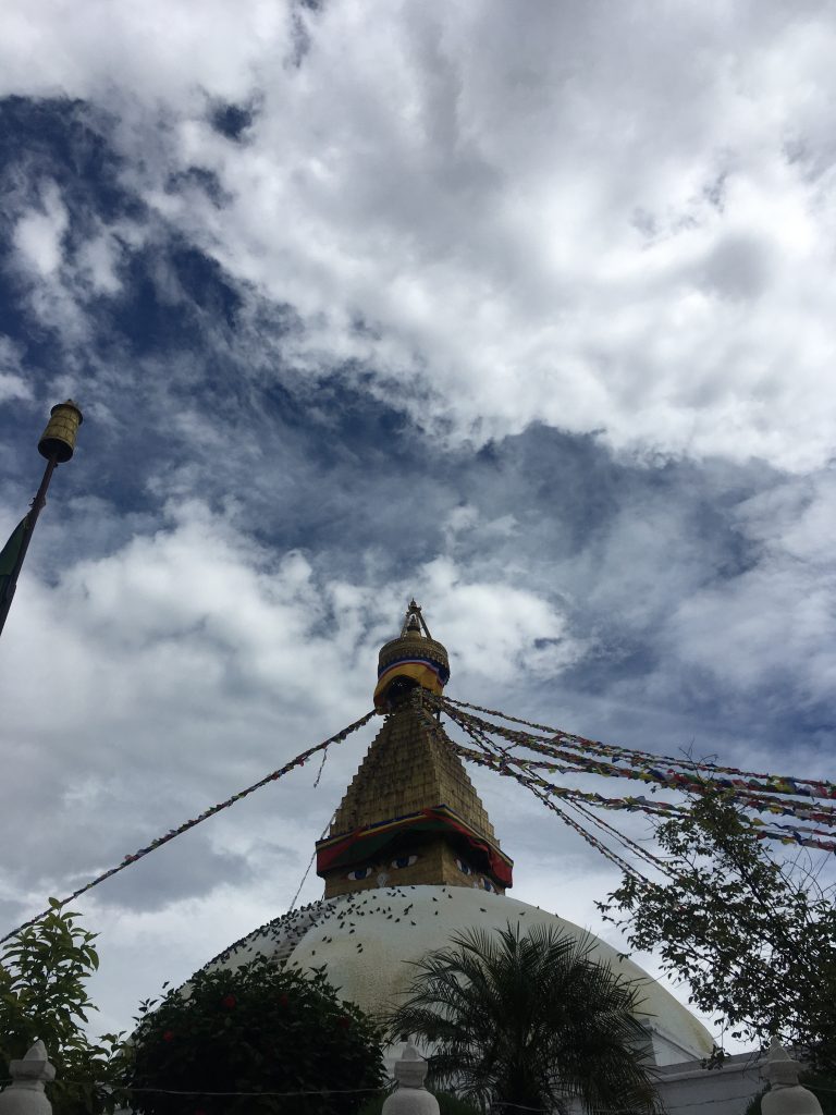 A large Bouddha stupa with eyes painted on it under a cloudy sky, adorned with colorful prayer flags extending from the spire.