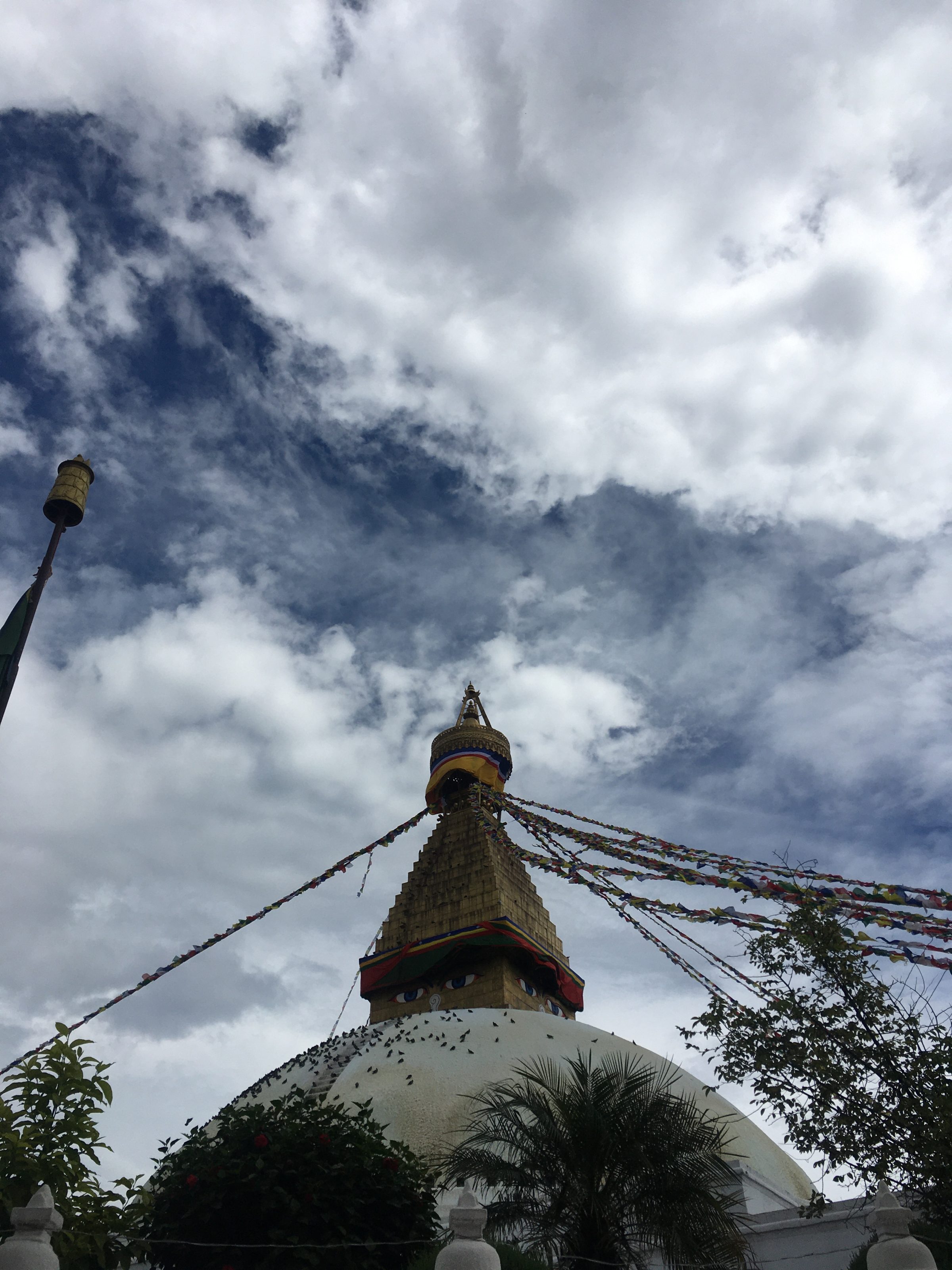 
A large Bouddha stupa with eyes painted on it under a cloudy sky, adorned with colorful prayer flags extending from the spire.