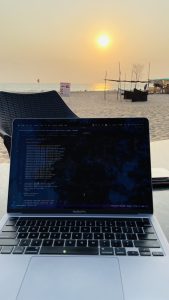 Laptop on a table with code on the screen, set against a beach backdrop at sunset. A wicker lounge chair, beach huts, and people are visible in the background.

