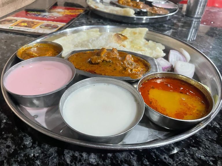 A metal tray with various Indian dishes, including bowls of pink and white desserts, curry, and dal. There are pieces of flatbread and sliced onions on the side.