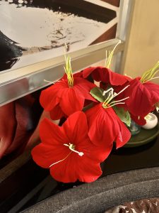 A cluster of vibrant red artificial flowers with prominent green stems and leaves, placed indoors near a framed picture with abstract patterns.