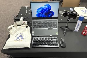 Photo of my workplace setup at WordCamp Asia 2025. There's a laptop on a laptop stand and a keyboard with a wireless mouse and phone used as a captioning software on a selfie stand.