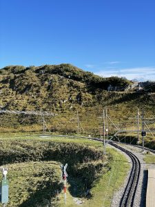 A scenic view of a railway track curving through a grassy hillside under a clear blue sky. The railway infrastructure includes poles and a crossing sign, and the area is surrounded by green vegetation and rolling hills