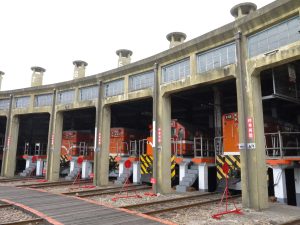 A row of train engines are parked inside a roundhouse with numbered sections. The engines are orange with a black and yellow diagonal stripe pattern on the front.