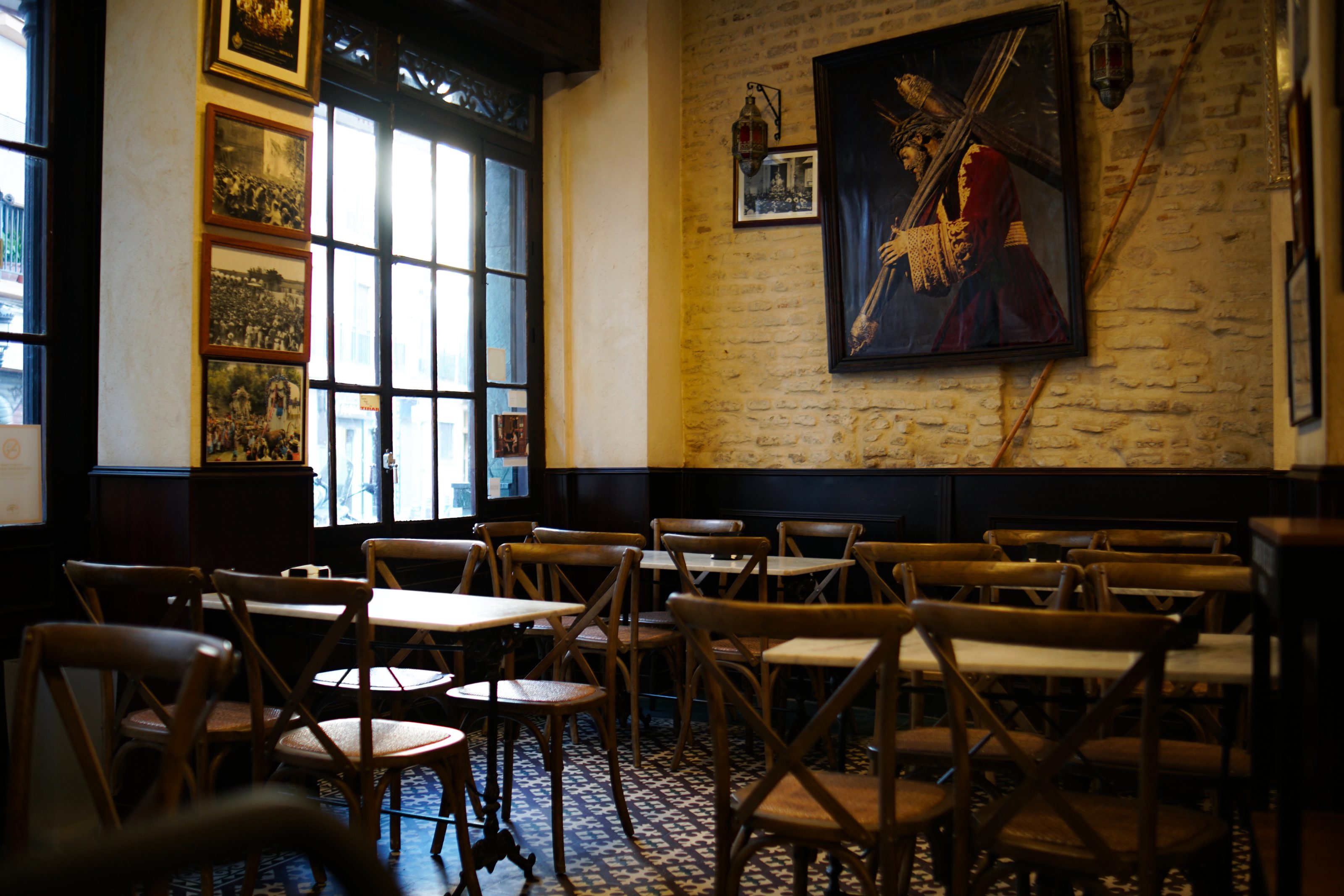 A cozy cafe interior with wooden chairs and tables arranged neatly on a patterned tile floor. Large windows allow natural light to illuminate the space. The decor features rustic elements, creating a warm and inviting atmosphere.