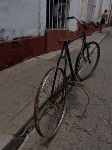 A rusted, old bicycle with no tires is parked on a cracked sidewalk against a weathered building with peeling paint. Cienfuegos, Cuba